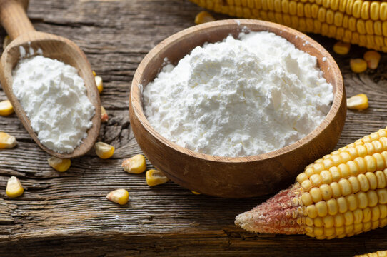Corn starch in bowl and spoon with dried corn groats, kernels on rustic table. Maize starch with ripe cobs and kernels