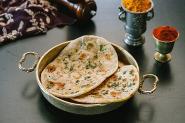 A platter showcases warm, freshly made Indian flatbreads, garnished with fresh herbs, accompanied by colorful spice containers, evoking the rich culinary heritage of India