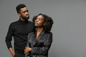 Elegant black couple in stylish outfits expressing love and confidence against a gray background, showcasing modern relationships in a contemporary setting