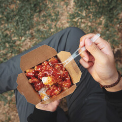 A person holds a takeout box filled with spicy fried chicken, accompanied by rice, in a box, enjoying food in a scenic natural environment