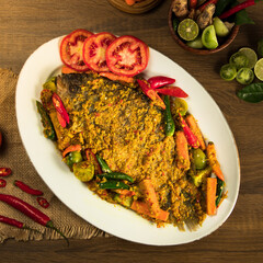 Indonesian traditional gurame pesmol, A beautifully presented grilled fish garnished with fresh tomatoes, chili peppers, and lime, surrounded by vibrant vegetables on a wooden table