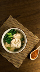 Wonton dumpling soup, A bowl of fresh vegetable and dumpling soup is placed on a bamboo mat with a side of spicy sauce, inviting warmth and flavor