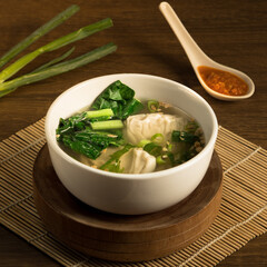 Wonton dumpling soup, A warm bowl filled with dumpling soup featuring fresh greens and a side of spicy sauce rests on a wooden table during a cozy meal
