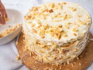 A hand sprinkling decorating crumbs over a pile of thin round layers spread with vanilla custard, a bowl of dough crumbs on a waffle towel in blurry background. Shortcrust puff pastry Napoleon cake.
