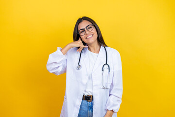 Young brunette doctor woman wearing stethoscope standing over isolated yellow background smiling doing phone gesture with hand and fingers like talking on the telephone
