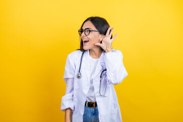 Young brunette doctor woman wearing stethoscope standing over isolated yellow background surprised with hand over ear listening an hearing to rumor or gossip