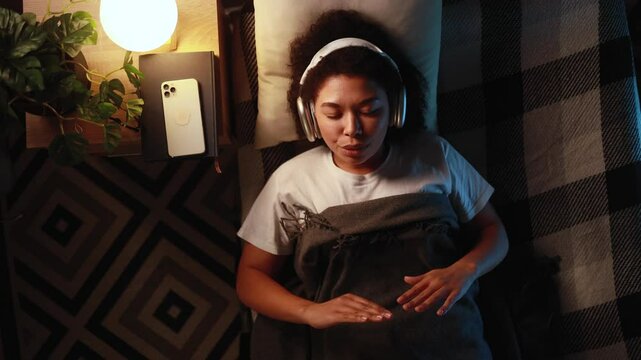 Top view of healthy mindful curly woman wearing headphones at bed listen calm music audio podcast doing yoga meditate relax exercise for mental balance feeling peace of mind before sleep at night