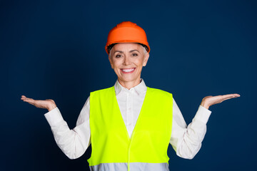 Confident senior businesswoman in bright uniform and orange helmet gesturing happily against a dark...