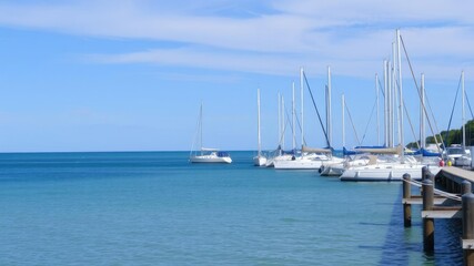 Fototapeta premium Sailboats docked along a scenic pier with calm sea waters and blue sky, waterfront, water sports, wave