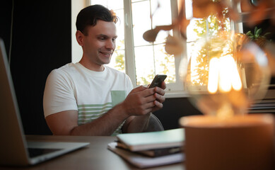 A man smiling while looking at his smartphone in a bright home office. He sits at desk with a laptop,notebooks, and warm light fixture.Sunlight streams through the window,creating relaxed atmosphere