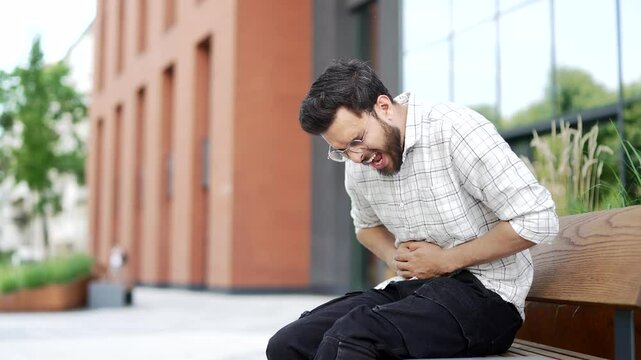 Sick man feels stomach pain while sitting on a bench on the street near a modern building. Upset male has heartburn, gastritis, bloating or poisoning. Businessman suffers from spasms and constipation