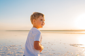 Young boy standing on a lakeshore during a sunny day, radiating happiness and tranquility, surrounded by a picturesque natural landscape.