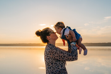 A loving mother lifting her happy baby during a warm sunset by a scenic lake, radiating joy and serenity.