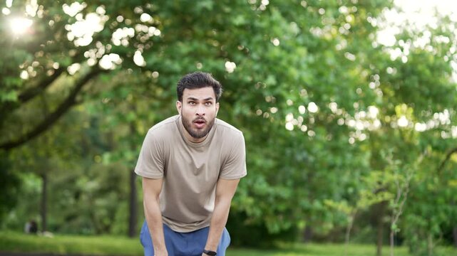 Adult runner has shortness of breath during a run in city park. Athlete feels tired. Sportsman rests his hands on his knees, taking a rest break, he's breathing heavily, wipes sweat from his forehead