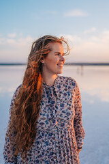 A young woman with long, windblown hair smiles peacefully by a calm lake during a golden sunset, radiating freedom, happiness, and natural beauty in a serene atmosphere.