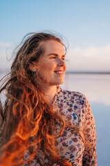 A young woman with long, windblown hair smiles peacefully by a calm lake during a golden sunset, radiating freedom, happiness, and natural beauty in a serene atmosphere.