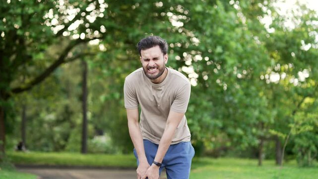 Young adult runner athlete with knee pain in an city park. Man massaging stretching, trauma injury while jogging outdoors. Fitness male sprain severe pain stretch pull. Leg muscle cramp calf sport