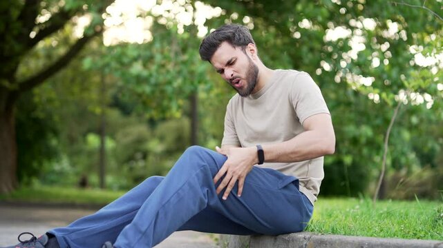 Upset athlete suffers from leg pain, calf muscle spasm during workout sitting on in an urban city park. Sporty male massaging knee cramps tendon at calves. Sprain severe pain stretch pull. Cramp calf