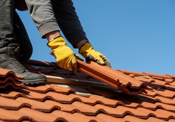 Person's hands wearing yellow gloves and black boots, installing a red tile on a red tiled roof with a zigzag pattern, against a clear blue sky.

