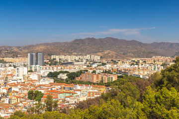 Obraz premium Malaga, seaside city in Andalusia, Spain, Europe. Panoramic view of a city nestled against mountains under a clear blue sky. Buildings, trees, and hills create a picturesque landscape.