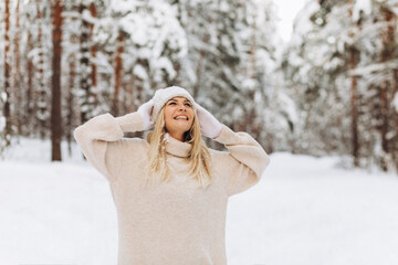 Winter portrait happy young woman in a sweater and knitted hat stands among the snowy forest