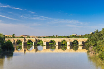 Fototapeta premium Roman bridge in Cordoba town in Spain. Ancient stone bridge spanning a calm river, reflecting the clear blue sky.