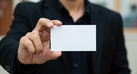 A close-up of a person holding a blank white business card at arm's length featuring a warm and inviting indoor setting the individual is wearing formal attire