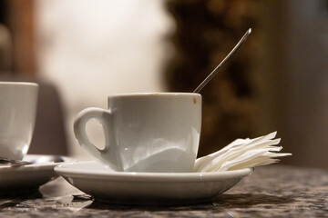 Minimalist coffee cup with a spoon and napkins on a polished granite surface in a softly lit cafe setting. Classic white espresso cup with a saucer and spoon on a granite table. Selective focus
