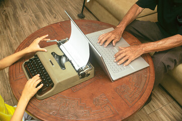 Senior writer using laptop next to typewriter and younger person using typewriter