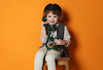 Cute little boy with old telephone on stool against orange background