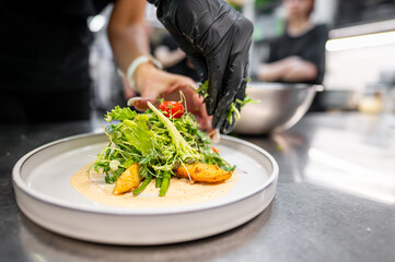 A chef in black gloves artfully garnishes a plate with fresh greens and vibrant vegetables, showcasing culinary creativity in a modern kitchen setting.