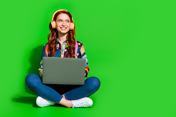 Bright young woman with red hair wearing headphones and holding a laptop on a vibrant green...