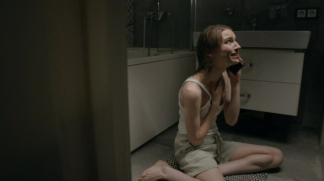 Impulsive Caucasian young woman wearing white tank top and light-colored shorts screaming and crying during intense conversation over phone on floor in bathroom
