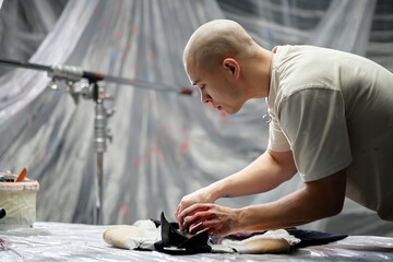 Side view of male fashion artist with buzz cut customizing clothes on table painting designer...