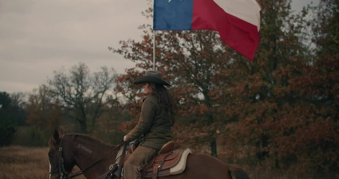 Woman Wearing Cowboy Hat Riding Galloping Horse Through Grassy Meadow While Holding Large Texas Flag With Cloudy Sunset Sky