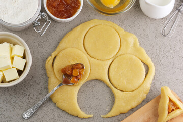 Preparation of a sweet pastry dough takes place in a cozy kitchen. Dough is rolled out and circular cutouts are combined with fruit filling and other baking ingredients.