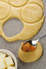 Preparation of a sweet pastry dough takes place in a cozy kitchen. Dough is rolled out and circular cutouts are combined with fruit filling and other baking ingredients.