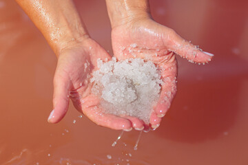 Close-up view of hands holding sparkling white salt crystals from the pink waters of a serene salt lake, with droplets of water reflecting sunlight and enhancing the natural beauty.