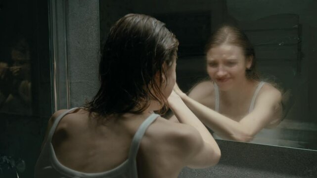 Young woman with depressive disorder frantically wiping away fog on mirror with both hands and crying in bathroom