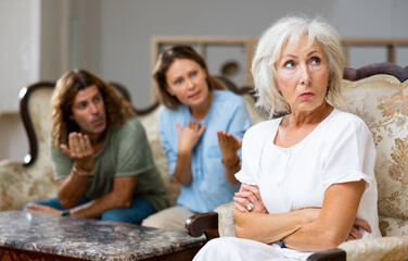 Angry mature female in armchair, keeping distance from the couple sitting on the couch at home