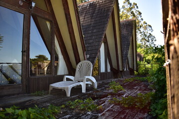 A-frame wooden cabin with a neglected terrace and aged plastic chairs, surrounded by lush vegetation. A nostalgic and rustic atmosphere in the heart of nature