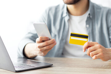 Shopping from home. Closeup of young guy holding credit card and smartphone, sitting in front of laptop, making online purchase at desk indoors. Young man buying goods on web, cropped view