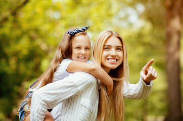 Fototapeta premium Look There. Mother Carrying Daughter Piggyback And Pointing Finger Spending Time Walking Together In Park Outdoor On Weekend