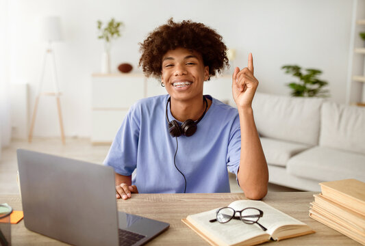 Happy black teenage student with headphones having creative idea near laptop, gesturing eureka at home. Afro teenager having aha moment while doing homework for online educational course