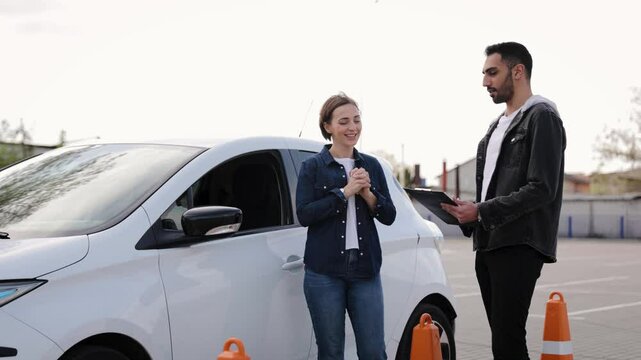 A driving instructor and his student stand by a white electric car, exchanging words during a driving lesson in a parking lot. Driving education with electric vehicle use