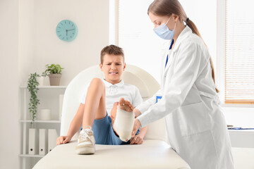 Fototapeta premium Female doctor examining little boy's injured leg on couch in clinic