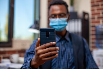 Businessman engaged in a videocall, holding his mobile device in brick wall office. Closeup of black male manager with face mask, grasping his smartphone and having virtual conversation with coworker.