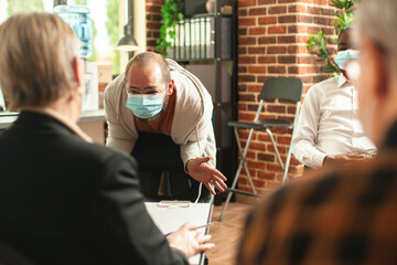 Caucasian angry man wearing face mask, sharing his frustrations with elderly female psychiatrist during group therapy. Distressed male person seeking help from retired therapist at counseling session.
