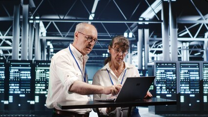 Senescent colleagues looking around high tech data center, using laptop to crosscheck disaster...