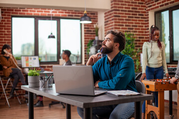 White business manager sitting at table with laptop, deep in thought, holding a pen to his chin. Male entrepreneur with a beard, reflecting on company projects as he looks around brick wall workplace.